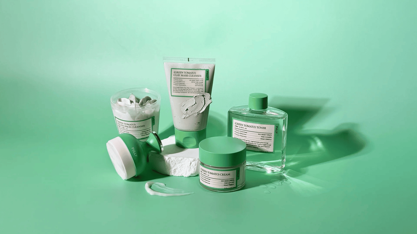 Gröna hudvårdsprodukter (cleanser, toner, kräm och mask) uppställda på ljusgrön bakgrund med kräm- och lertexturer i fokus.