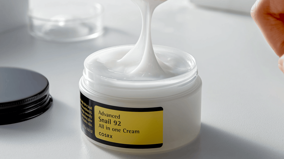 En öppen burk av COSRX Advanced Snail 92 All in one Cream med tjock, vit kräm som lyfts med en spatel, på en vit yta.