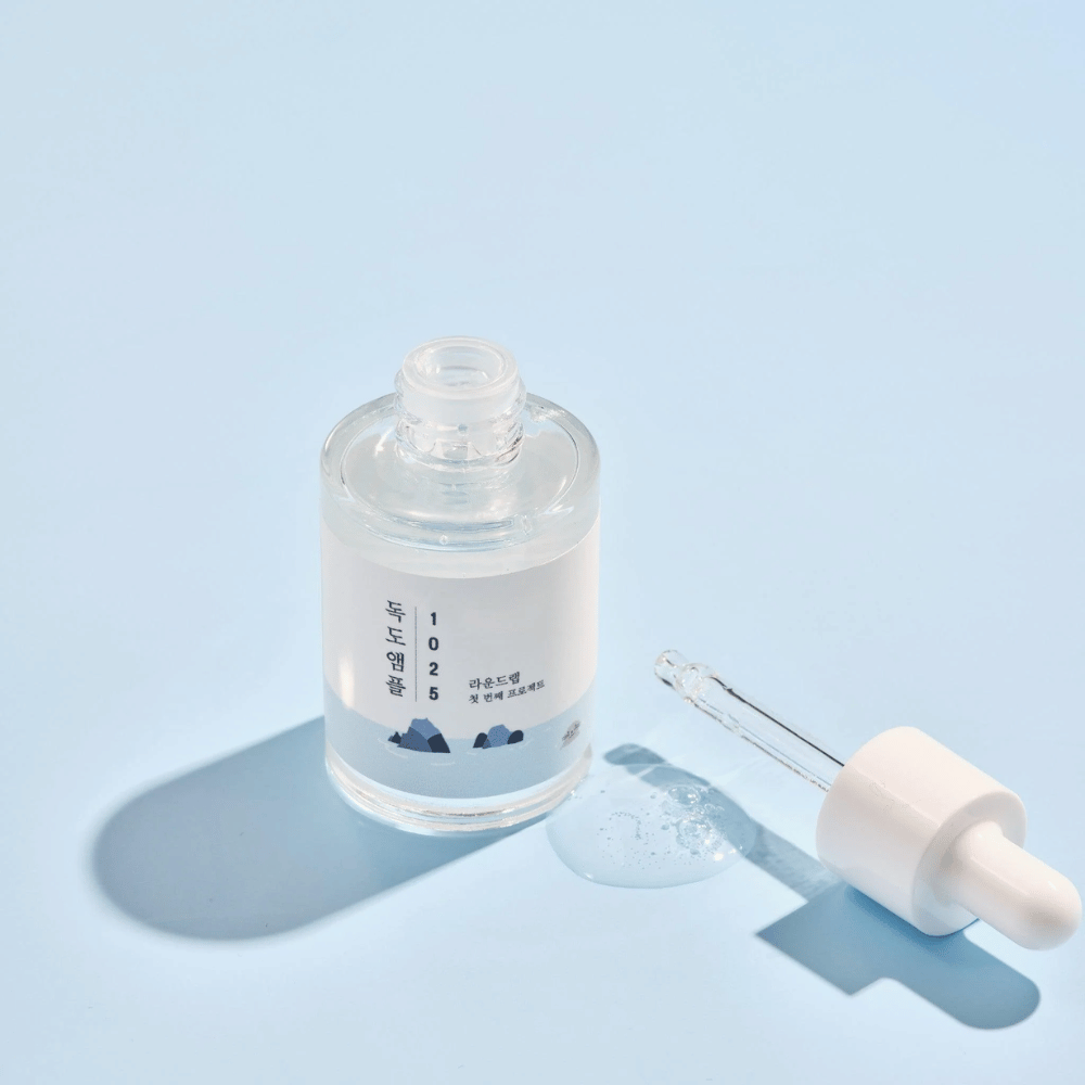En åpen flaske av 1025 Dokdo Ampoule med en dråpeteller ved siden av, mot en blå bakgrunn som skaper en myk skygge.