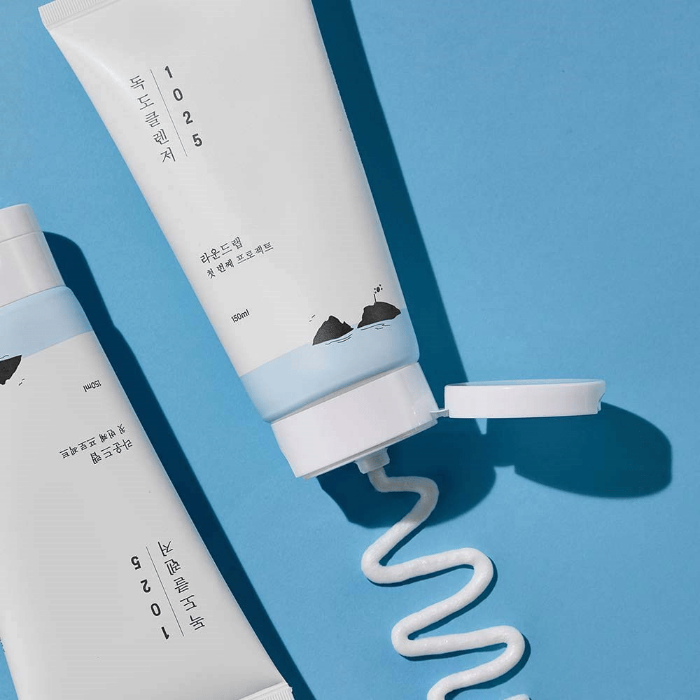 To tuber av 1025 Dokdo Cleanser på blå bakgrunn, hvorav en med åpen kork og en bølgete streng rensing presset ut.