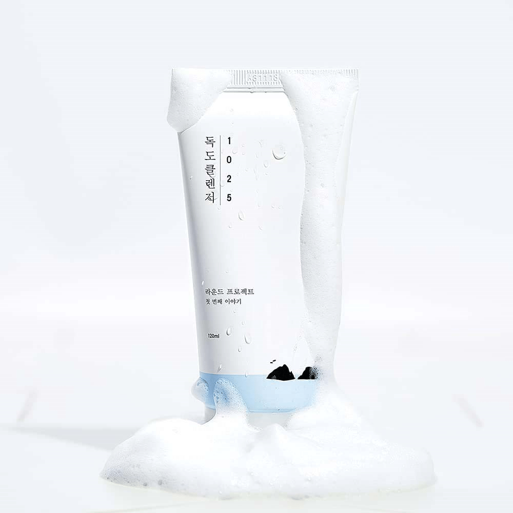 1025 Dokdo Cleanser i hvit tube dekket av skum, med vannperler og skum rundt bunnen, frilagt mot lys bakgrunn.