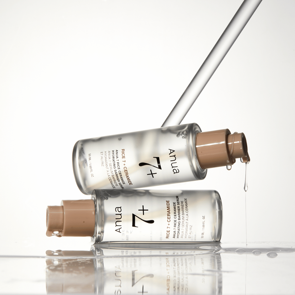 Bildet viser to gjennomsiktige flasker av "Anua 77 Rice + Ceramide Hydrating Barrier Serum" med dråper som renner fra en pipette. Serumet, som er beriket med ris og ceramider, er ment å fukte og styrke hudens barriere. Flaskene er elegant designet, noe som forsterker produktets premiumfølelse.