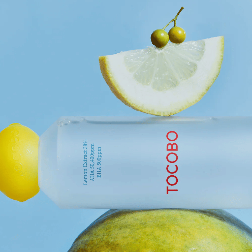 Flaske med TOCOBO AHA BHA Lemon Toner, dekorert med et lokk i form av en sitron. En sitronskive og små grønne bær balanserer oppå flasken mot en blå bakgrunn.