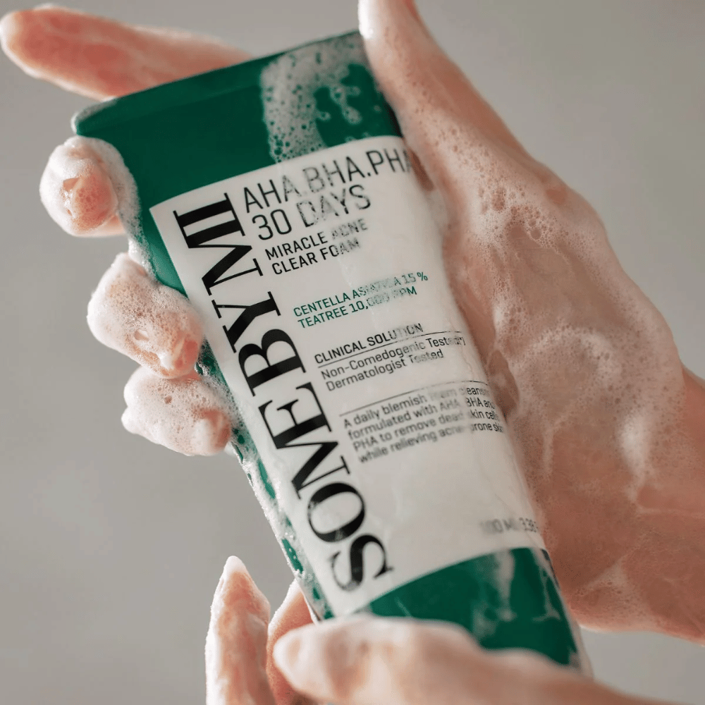 En person holder en tube av "SOME BY MI AHA, BHA, PHA 30 Days Miracle Acne Clear Foam" med skummende innhold over hendene. Produktet er ment for daglig bruk for å bekjempe akne og rense porene med sine aktive ingredienser som centella, tea tree olje og syrer som AHA, BHA og PHA.