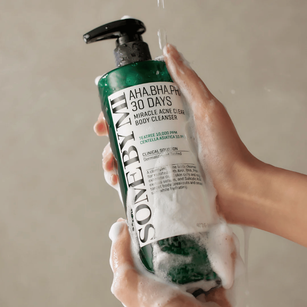 En person holder i en flaske SOME BY MI AHA.BHA.PHA 30 Days Miracle Acne Clear Body Cleanser. Flasken er grønn med en hvit etikett og er dekket av skum fra rengjøringsmiddelet. Personen vasker seg med produktet mens vann renner over hendene.