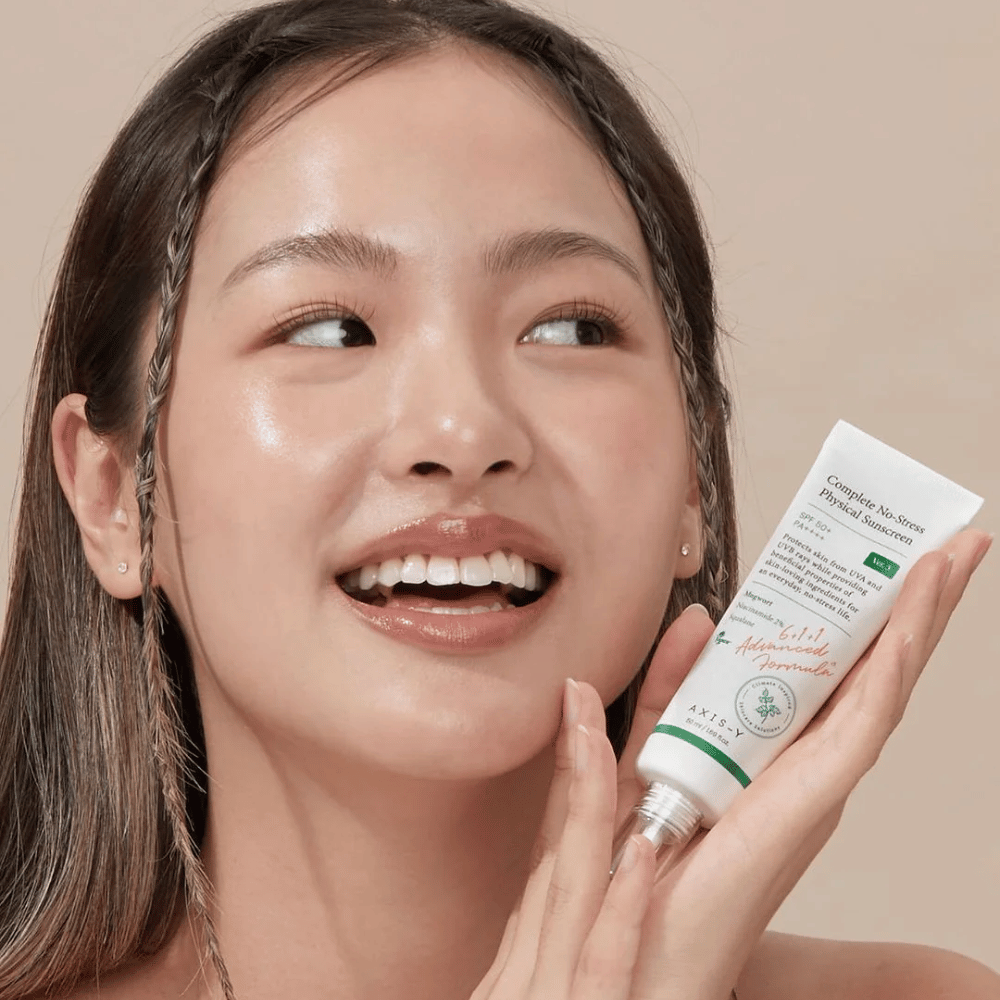 Bildet viser en ung kvinne som smiler bredt og holder en tube med AXIS-Y Complete No-Stress Physical Sunscreen. Hun har et strålende og naturlig utseende med minimal sminke, som fremhever produktet hun presenterer.