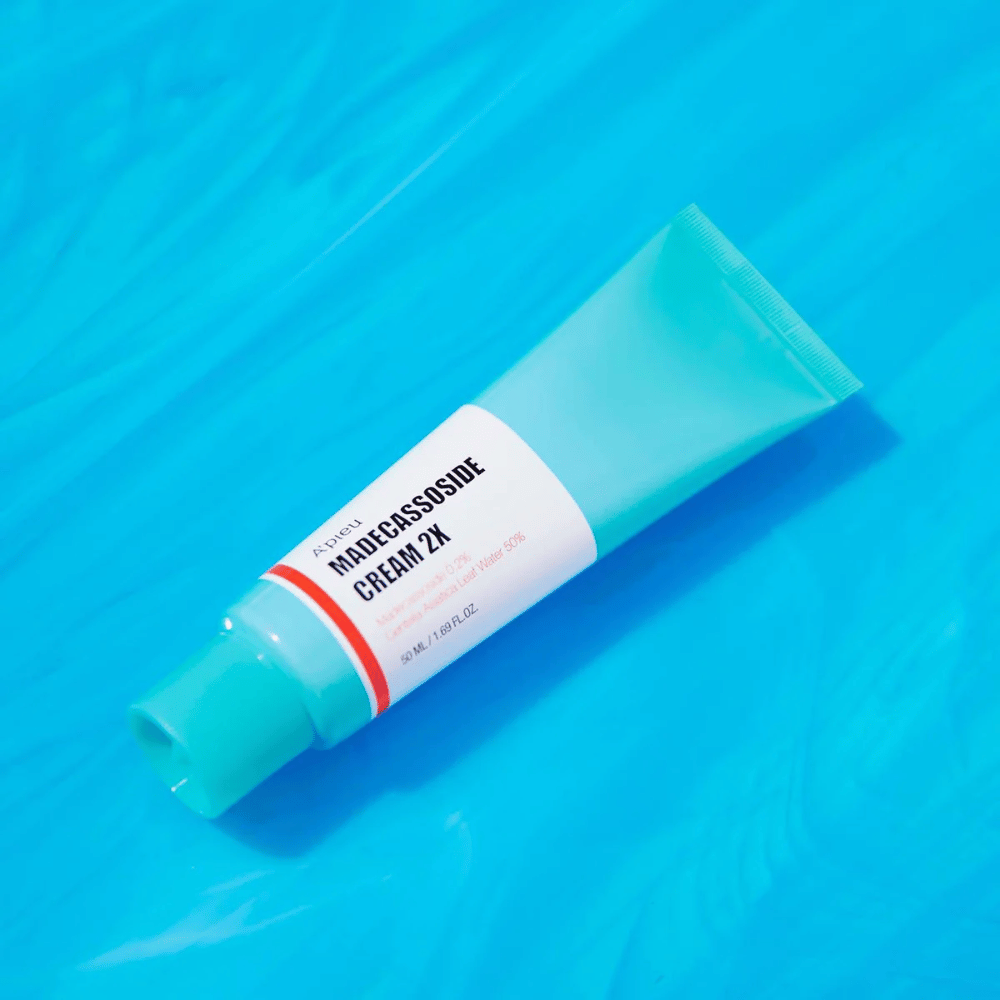 En tube A'PIEU Madecassoside Cream 2X ligger diagonalt over en blå bakgrunn med vannlignende rynker. Tuben har en hvit kropp med en lys turkis kork og en rød til oransje farget etikett der produktnavnet og den aktive ingrediensen Madecassoside 2% er tydelig skrevet ut. På etiketten står også mengden som er 50 ml / 1.8 fl oz. Den lyse og fargerike presentasjonen gir en frisk og energisk følelse.