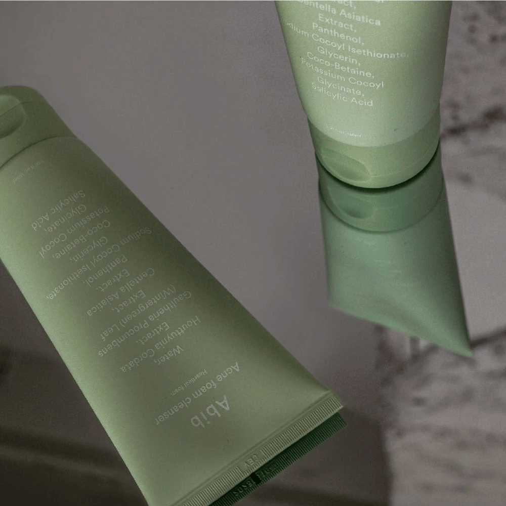Grønn tube av Abib Acne Foam Cleanser Heartleaf liggende på et speilbord med produktets forside og tekst speilet. Ingredienser som salisylsyre, centella og hjertebladekstrakt vises tydelig på emballasjen.