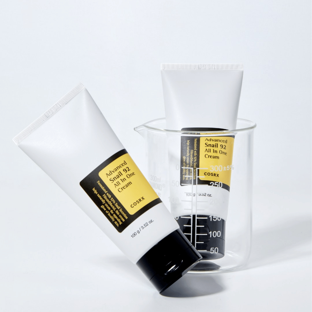 Et rør av COSRX Advanced Snail 92 All in One Cream liggende ved siden av en opp-ned gjennomsiktig målesylinder på en hvit bakgrunn.