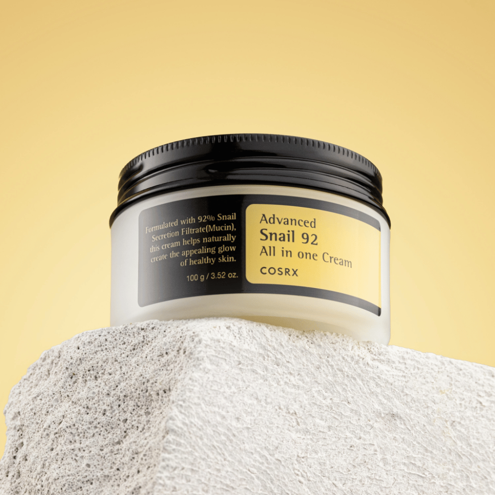 En krukke av COSRX Advanced Snail 92 All in one Cream plassert på en steinete overflate med en gul bakgrunn.