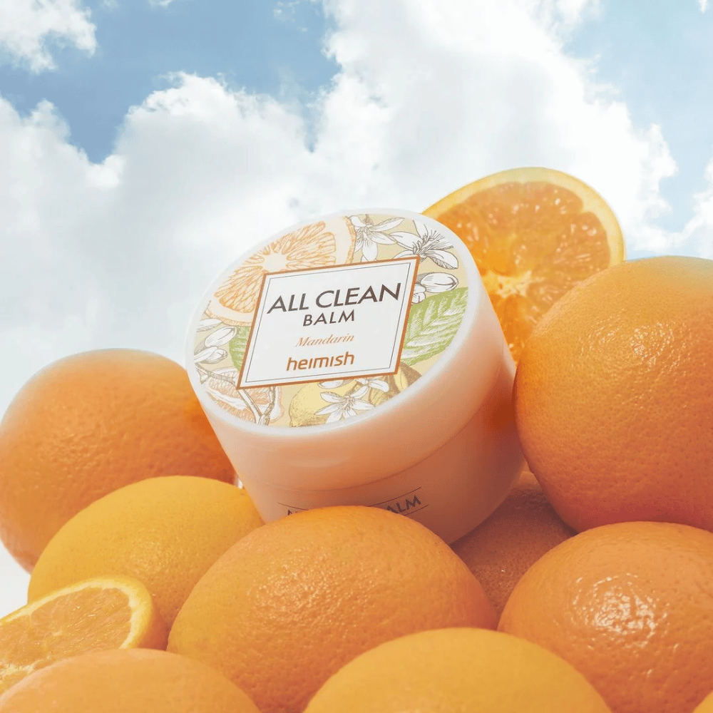 Krukke av Heimish All Clean Balm Mandarin plassert på en haug med appelsiner, med en blå himmel i bakgrunnen.