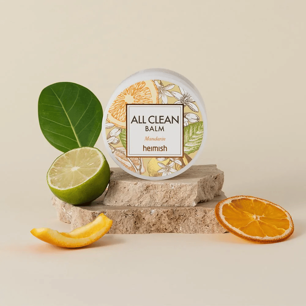 Krukke av Heimish All Clean Balm Mandarin plassert på steiner, omgitt av mandarinskall og sitrusblader, foran en lys bakgrunn.