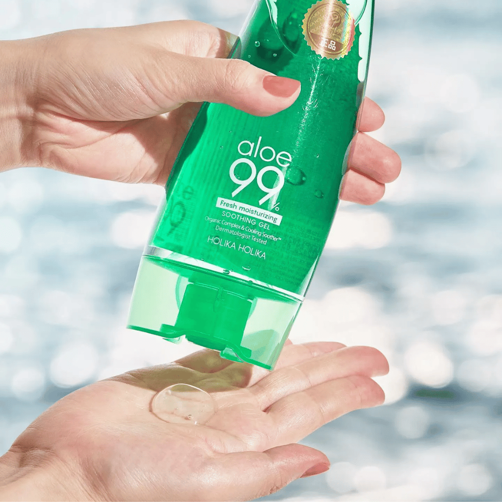 En hånd holder en grønn tube med Holika Holika Aloe 99% Soothing Gel, mens en annen hånd fanger en klikk gel som dispenseres. Bakgrunnen er uskarp og viser et vannaktig, glitrende mønster.
