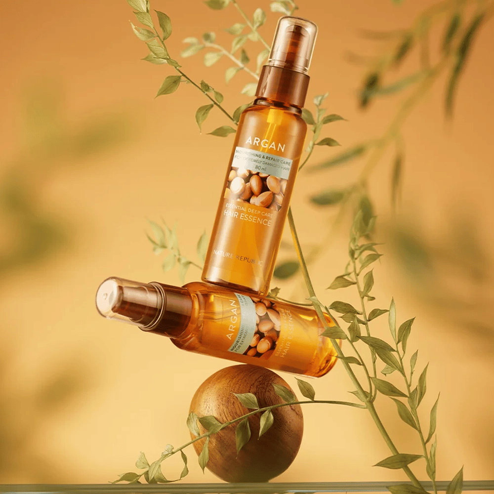To flasker Nature Republic Argan Essential Moisture Hair Oil plassert blant grønne blader – silkemyk glans for tørt hår.