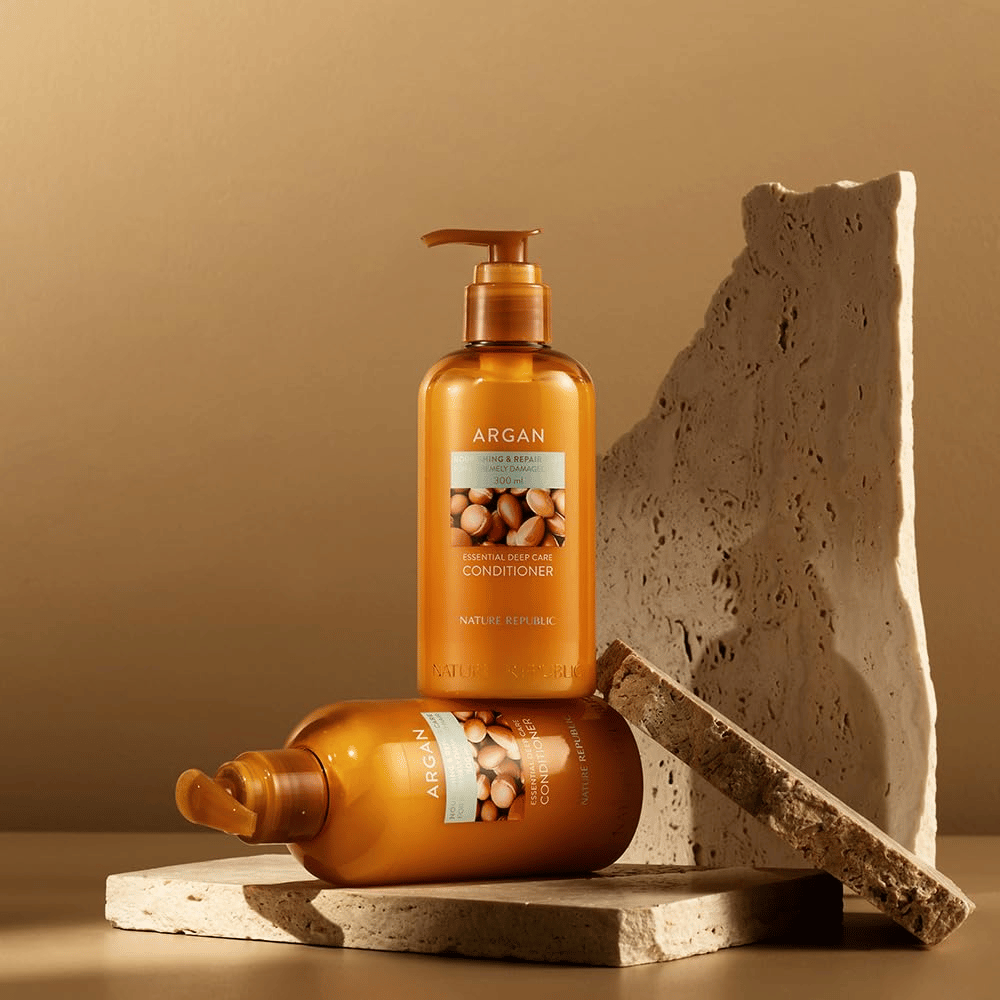 To flasker av Nature Republic Argan Essential Moisture Repairing Conditioner på steinunderlag i varm tone.