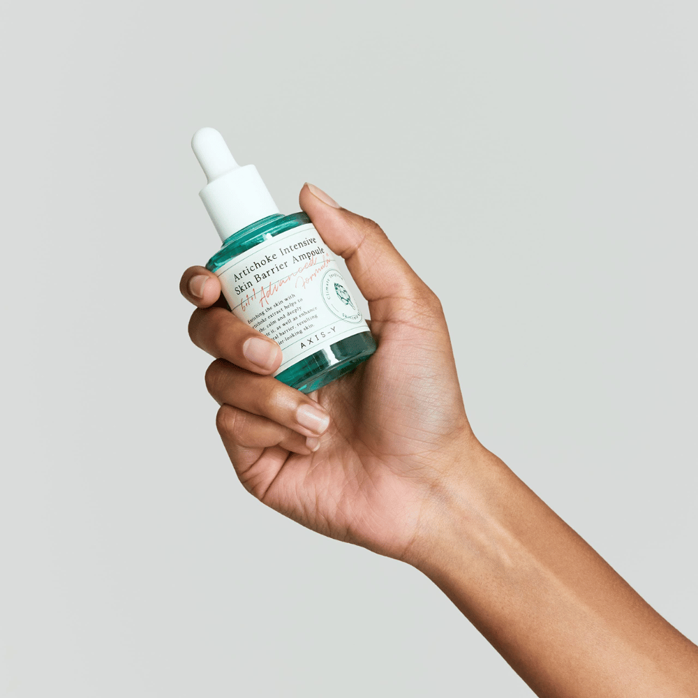 En hånd holder en flaske hudpleieserum med etiketten "Artichoke Intensive Skin Barrier Ampoule.