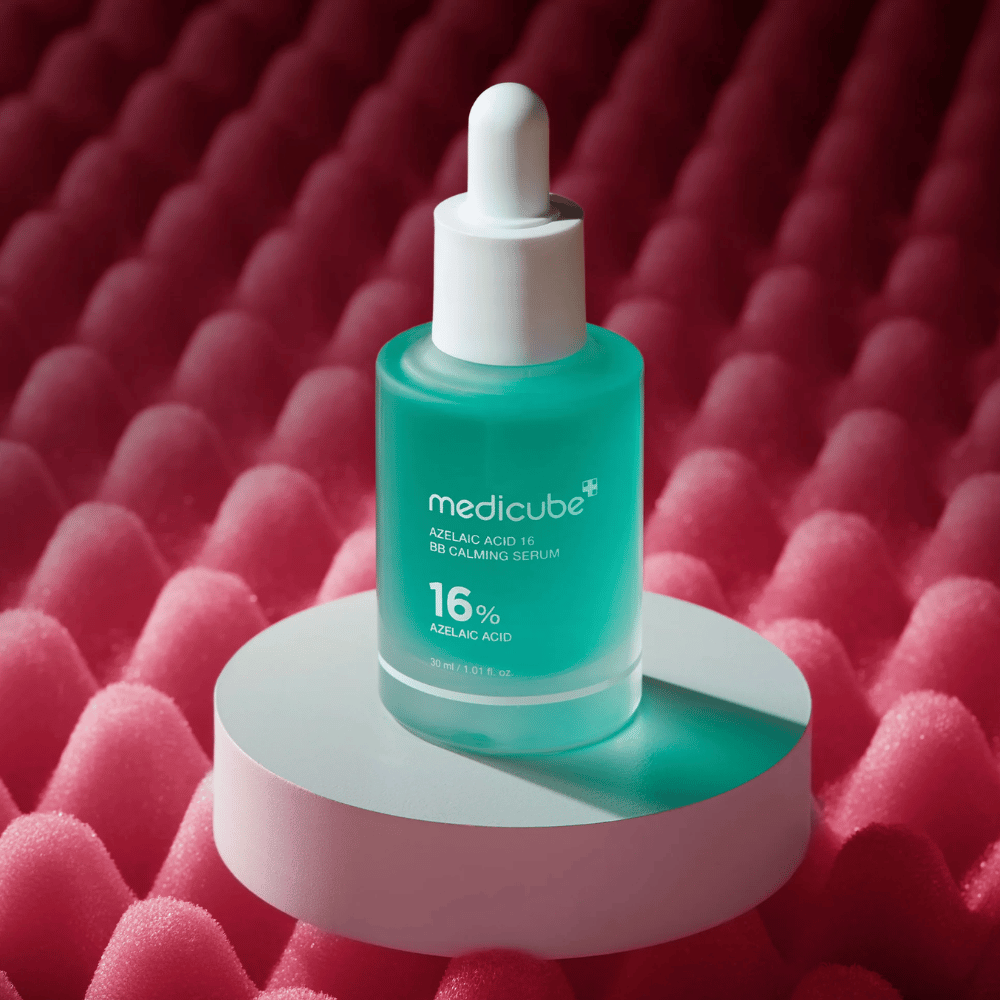 Produktbilde av Medicube Azelaic Acid 16 BB Calming Serum plassert på hvit podiumoverflate med rosa strukturert bakgrunn som fremhever serumets beroligende og hudforbedrende profil.