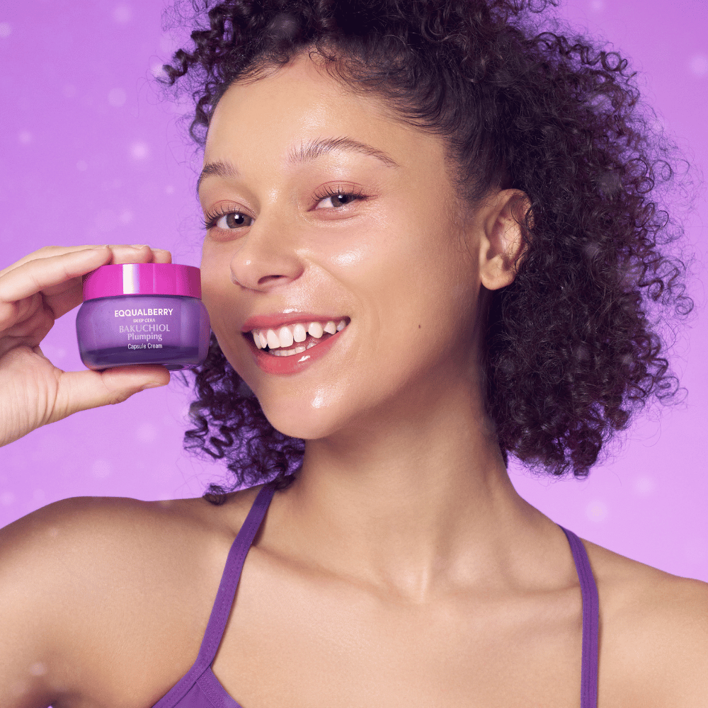Kvinne med glødende hud holder EQQUALBERRY Bakuchiol Plumping Capsule Cream og smiler foran en lilla bakgrunn.
