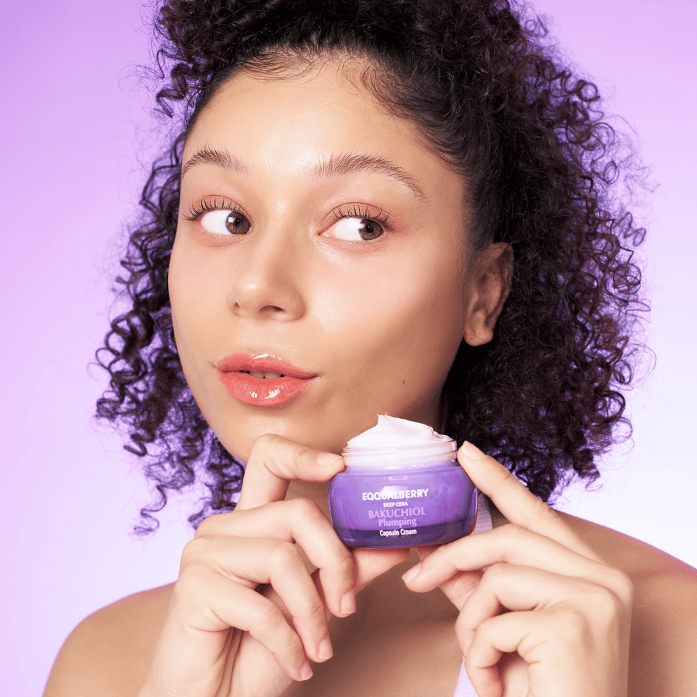 Kvinne holder EQQUALBERRY Bakuchiol Plumping Capsule Cream i hånden og viser frem sin fuktige, glødende hud mot en lilla bakgrunn.