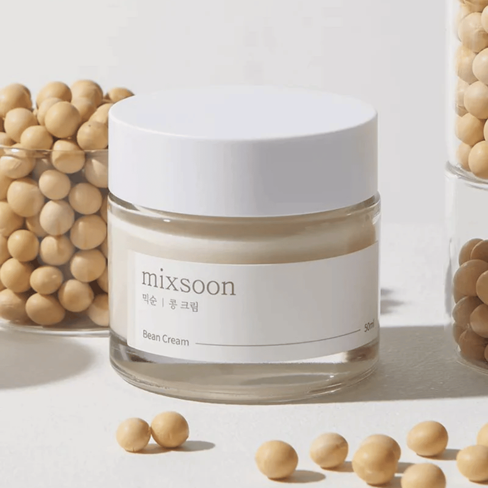 En gjennomsiktig boks med hvit etikett og lokk, merket "mixsoon 팥 크림" og "Bean Cream" med en angitt volum på "50ml". Boksen inneholder en lysegul krem og er plassert på en lys overflate. Ved siden av boksen er det en glassbeholder full av lysebrune bønner som er spilt ut på overflaten, noe som antyder at kremen inneholder bønneekstrakt. Bakgrunnen og belysningen gir en varm og naturlig atmosfære.