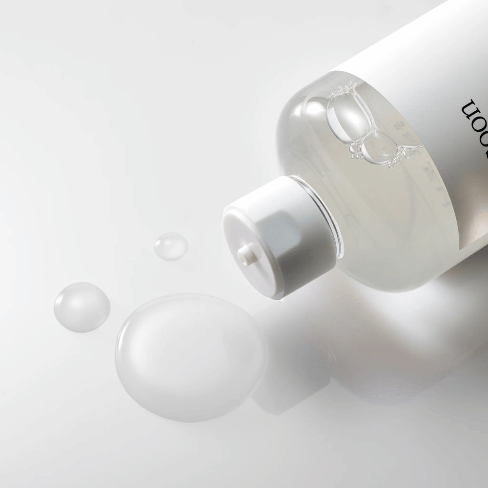 En gjennomsiktig flaske ligger på siden på en blank overflate med en lett toner som sakte renner ut og danner dråper og en større dam på overflaten. Flasken har en hvit etikett og en hvit pumpemekanisme. Belysningen er myk og skaper en ren og beroligende atmosfære, mens tonerns lettflytende konsistens og klarhet fremheves gjennom hvordan den sprer seg på overflaten.