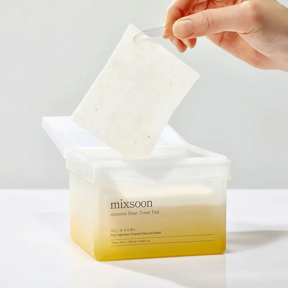 En hånd holder en pinsett og løfter opp en Mixsoon Bean Toner Pad fra en firkantet, gjennomsiktig beholder med en gulaktig tone. Beholderen inneholder 70 toner pads og har tekst som beskriver produktet.