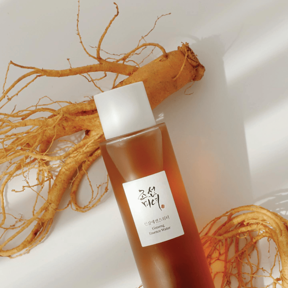 En brun flaske Beauty of Joseon Ginseng Essence Water med hvit kork ligger ved siden av en fersk ginsengrot på hvit bakgrunn.