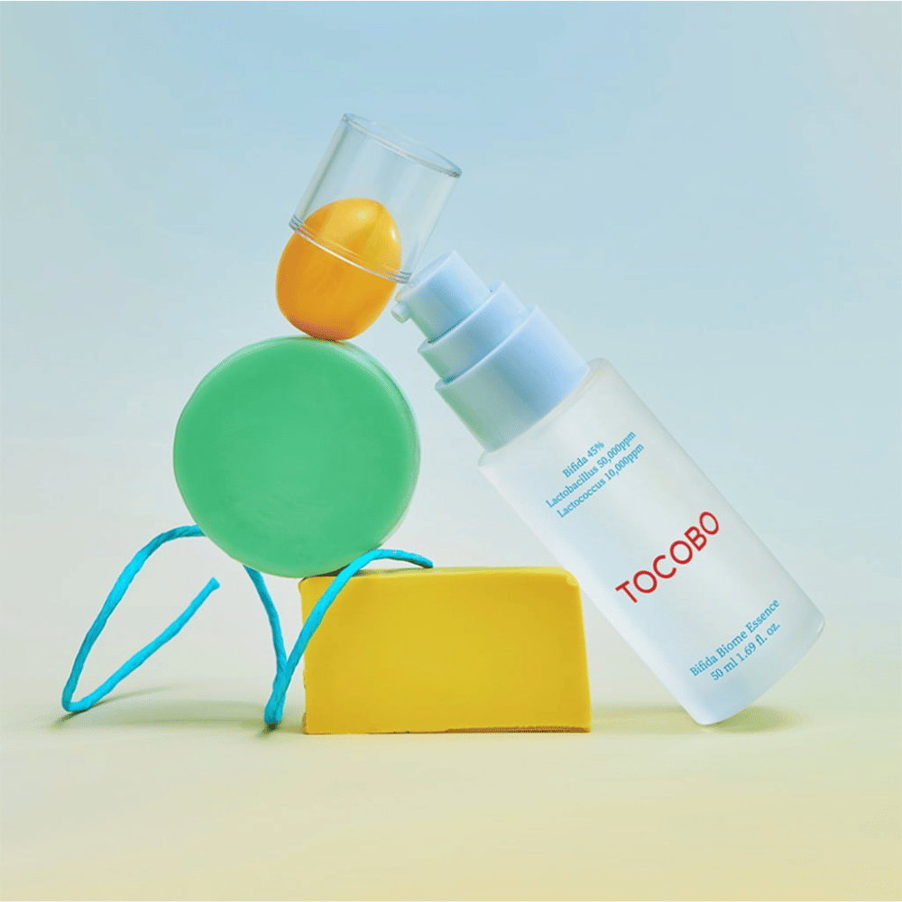 TOCOBO Bifida Biome Essence i frostet pumpeflaske med rød logo, plassert mot fargerik bakgrunn med grønne, gule og oransje geometriske former. Tekst på flasken angir Bifida 5 %, Lactobacillus 3 000 ppm og Lactococcus 1 000 ppm.