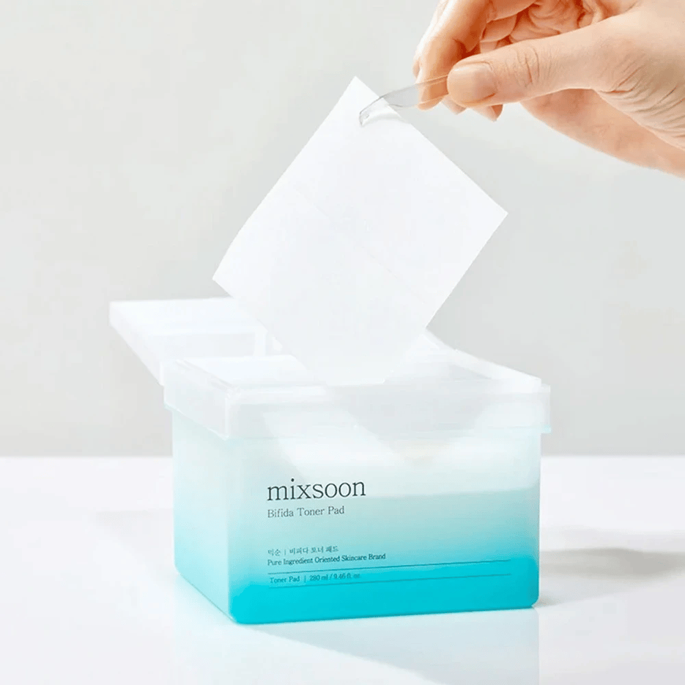 En gjennomsiktig beholder med en blå og hvit gradient som inneholder Bifida toner pads. En hånd tar en toner pad fra beholderen, som er merket med "mixsoon Bifida Toner Pad" og rommer 280 ml (9,46 fl. oz.) toner pads beregnet for hudpleie.