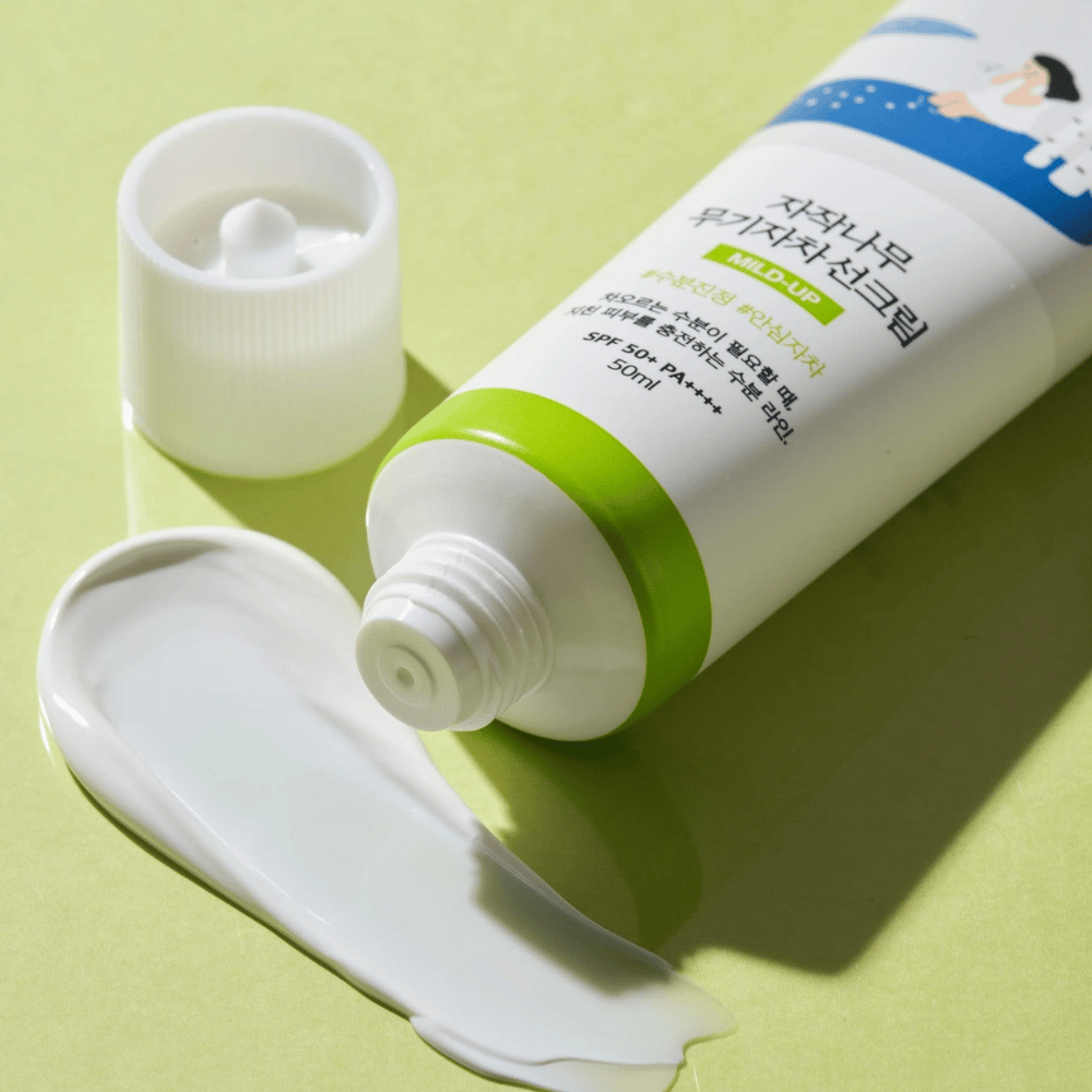 Nærbilde av Birch Juice Moisturizing Mild Up Sunscreen med skrudd av kork, hvit solkrem smurt ut på lys grønn bakgrunn, SPF 50+ PA++++, 50 ml.