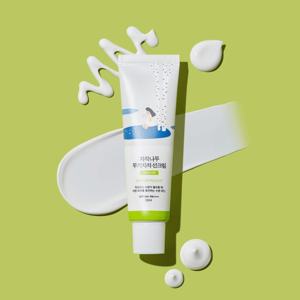 Birch Juice Moisturizing Mild Up Sunscreen – hvit tube med grønn base på grønn bakgrunn, omgitt av smurt ut solkrem og dråper, SPF 50+ PA++++, 50 ml.