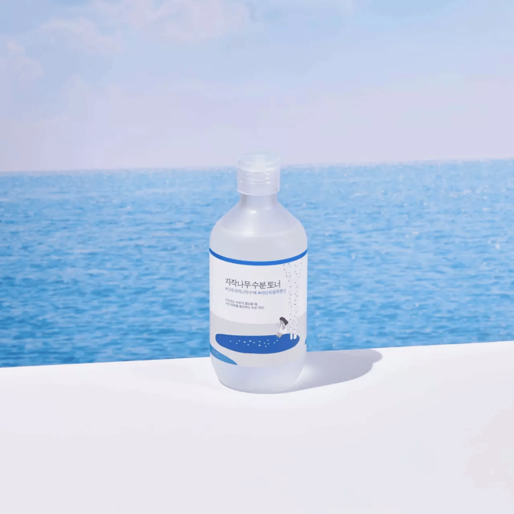 Flaske av ROUND LAB Birch Juice Moisturizing Toner står på en hvit overflate med hav og blå himmel i bakgrunnen
