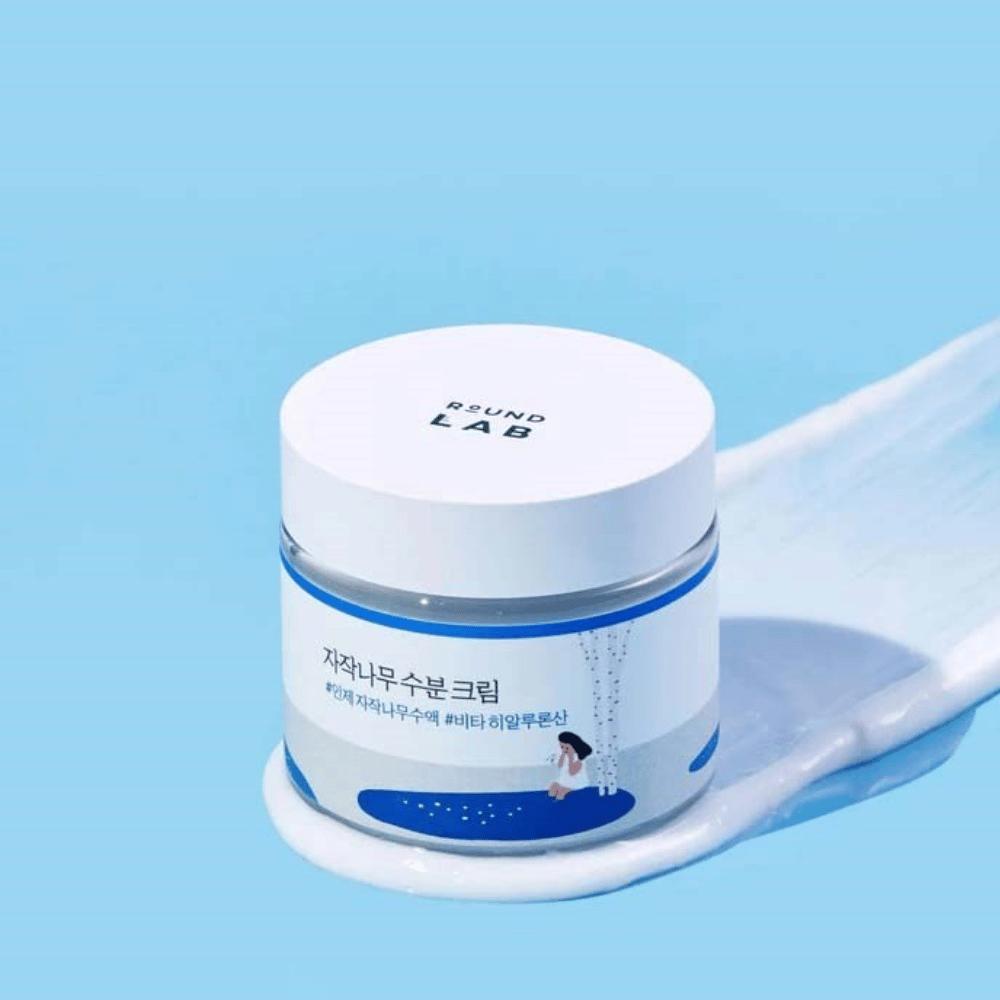 Round Lab Birch Juice Moisturizing Cream i blåtonet boks med hvitt lokk, stående i et lag av krem mot blå bakgrunn.