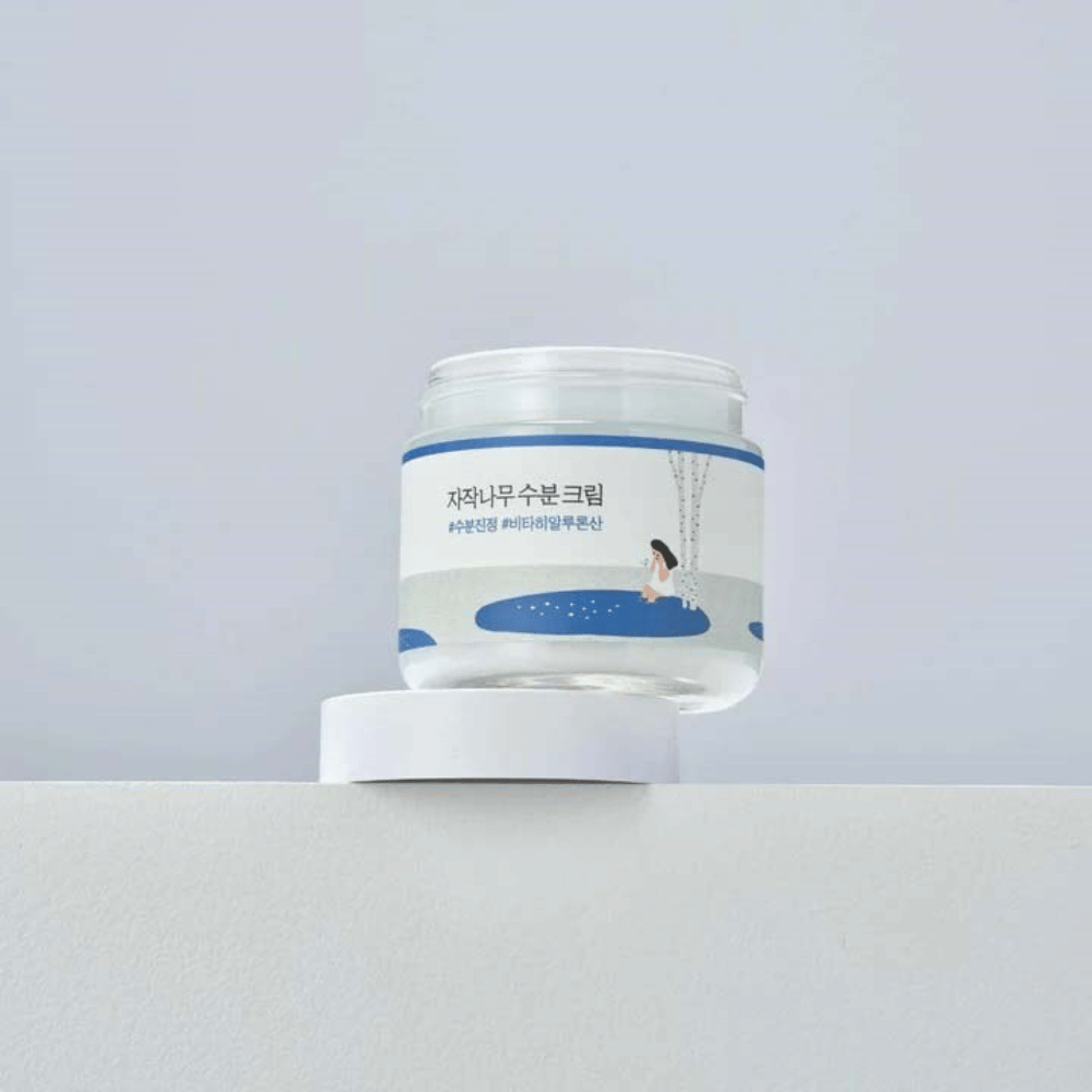 Round Lab Birch Juice Moisturizing Cream i gjennomsiktig boks med hvit etikett og blå illustrasjon av kvinne ved bjørk, stående på hvit overflate mot lys bakgrunn.