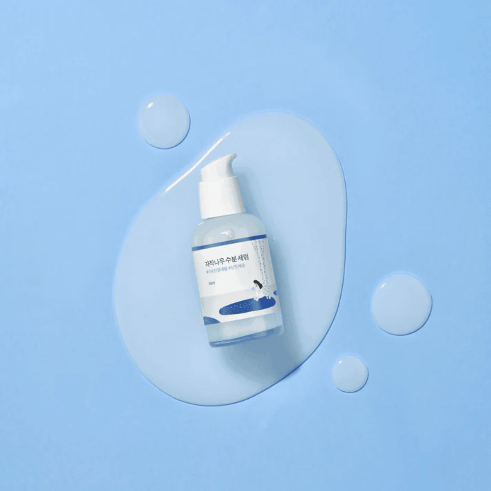 Round Lab Birch Juice Moisturizing Serum i gjennomsiktig pumpeflaske plassert på en blå bakgrunn med dråper og utøst serum.