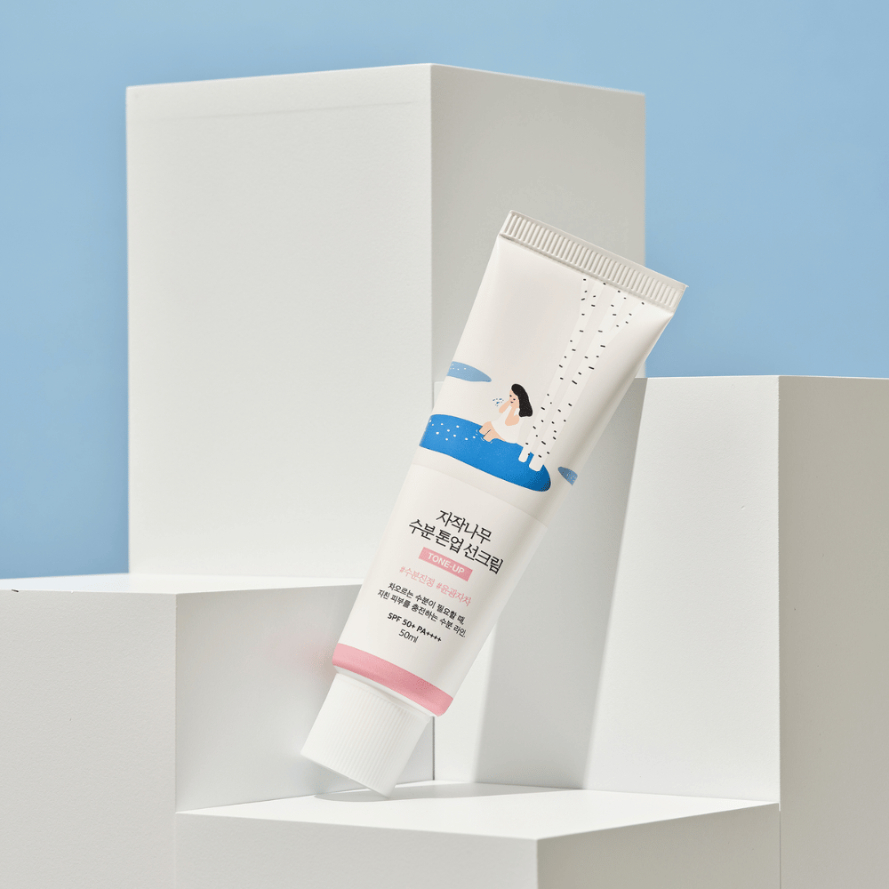 En tube med Birch Juice Moisturizing Tone Up Sunscreen plassert på hvite kuber mot en lys blå bakgrunn. Tuben er hvit med en illustrasjon av en kvinne ved en bjørk og vann. Teksten på koreansk angir at produktet har SPF 50+ PA++++ og inneholder 50 ml.