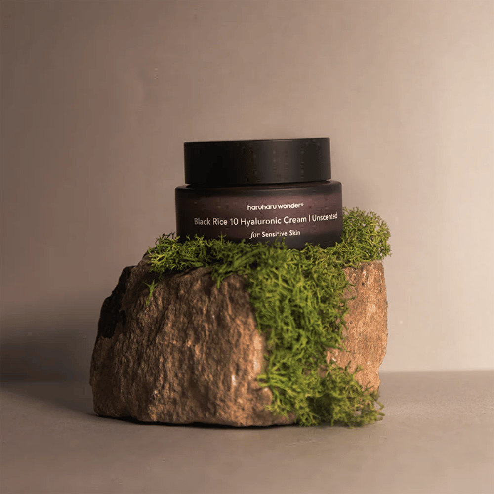 Bildet viser en krukke hudkrem merket "haruharu wonder Black Rice 10 Hyaluronic Cream Unscented for Sensitive Skin". Krukken er svart og står på en stor stein dekket med lysegrønn mose, noe som skaper en naturlig og jordnær følelse.