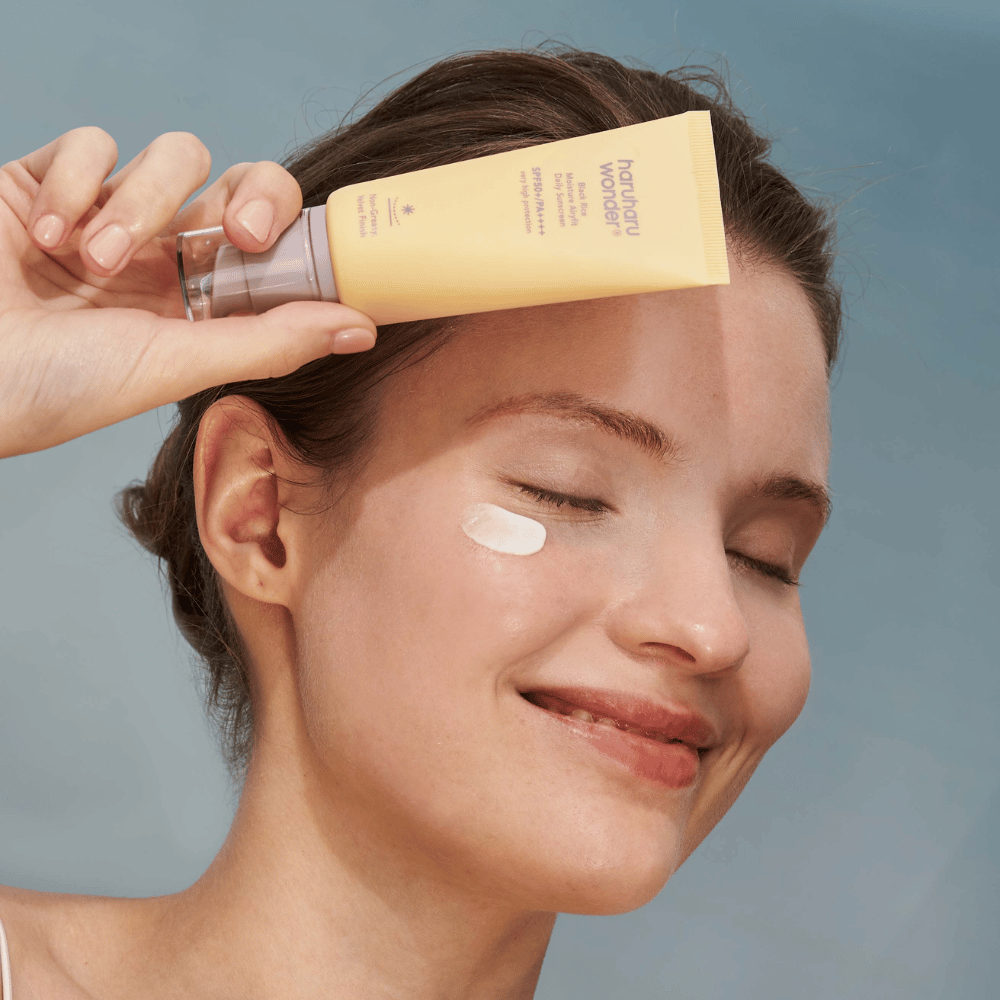 En smilende kvinne påfører Black Rice Moisture Airyfit Daily Sunscreen SPF 50+ PA++++ fra Haruharu Wonder på ansiktet sitt. Hun holder en gul tube med solkrem og har en klatt av produktet på kinnet. Bakgrunnen er lyseblå.