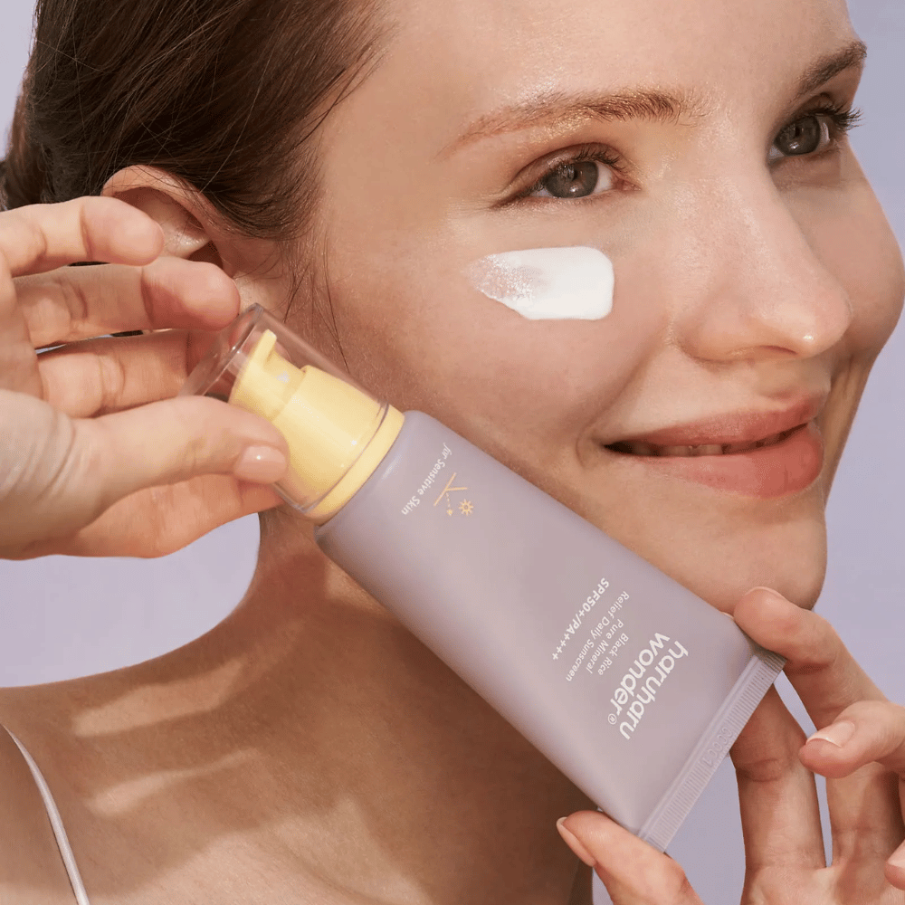 En smilende kvinne påfører Haruharu Wonder Black Rice Pure Mineral Relief Daily Sunscreen SPF 50+ PA++++ på ansiktet sitt. Hun holder en lilla tube med solkrem og har en klatt av produktet på kinnet. Bakgrunnen er lyse lilla.