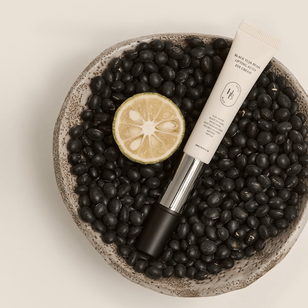 Black Yuja Bean Lifting Auto Eye Cream liggende i en bolle fylt med svarte bønner og en halvert sitrusfrukt – en visuell representasjon av produktets naturlige ingredienser.