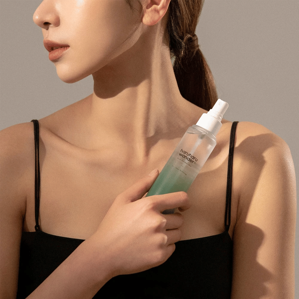 Bilde av HaruHaru Wonder Black Bamboo Mist som holdes av en kvinne. Produktet er i en gjennomsiktig sprayflaske med en grønn tone og en hvit pumpe. Kvinnen har et avslappet utseende, har på seg en svart topp og fokuserer på å bruke misten for å gi huden fuktighet og glød. Flasken er en del av en hudpleierutine.