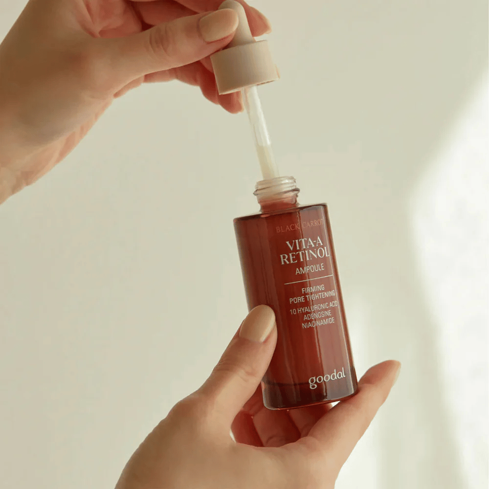 En hånd holder en flaske med Goodal Black Carrot Vita-A Retinol Firming Ampoule. Flaskens etikett viser produktets navn og egenskaper, inkludert "Firming Pore Tightening" og ingredienser som 10% hyaluronsyre, adenosin og niacinamid. Pipetten er i fargen beige.