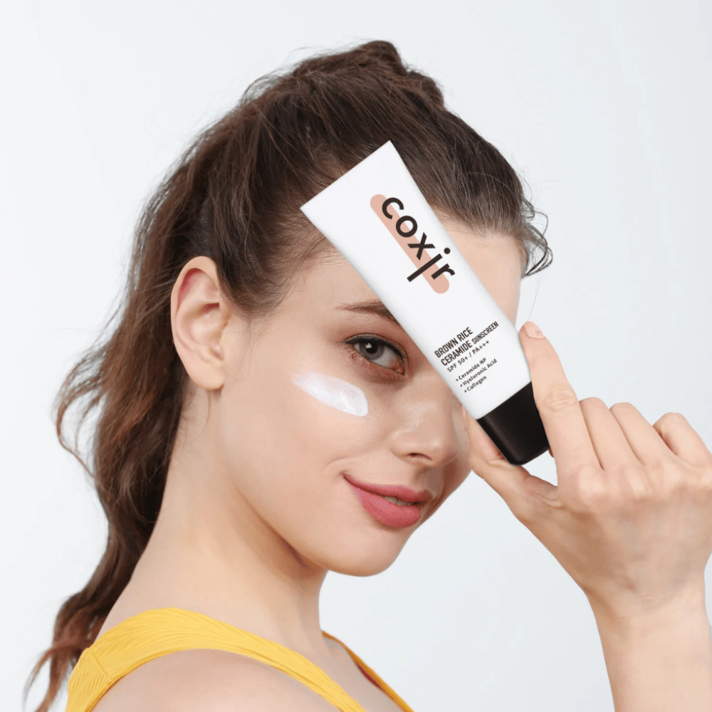Kvinne med solkrem påført på kinnet holder opp en tube Coxir Brown Rice Ceramide Sunscreen, SPF 50+ PA+++, mot pannen og smiler lett.