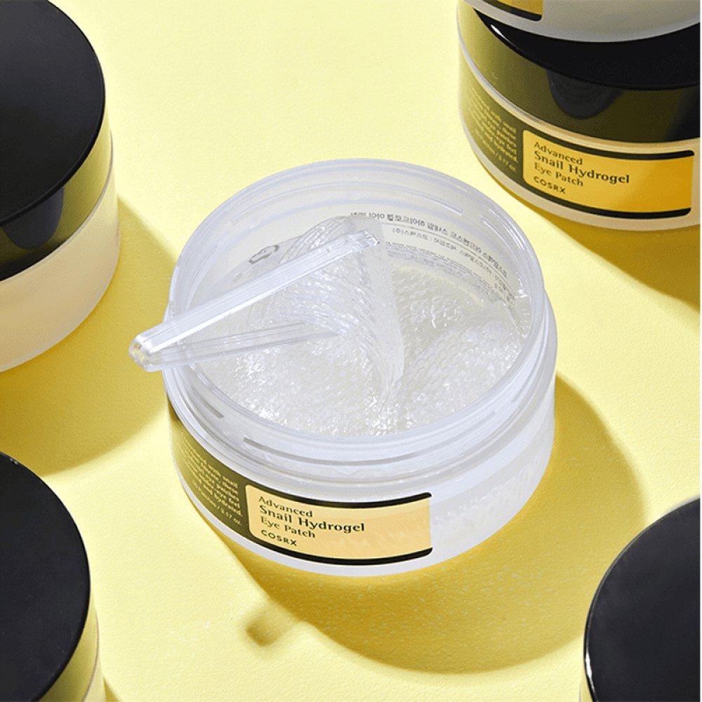 En åpen krukke COSRX Advanced Snail Hydrogel Eye Patch med gjennomsiktige øyenlapper og en liten spatel, omgitt av flere lukkede krukker på gul bakgrunn.