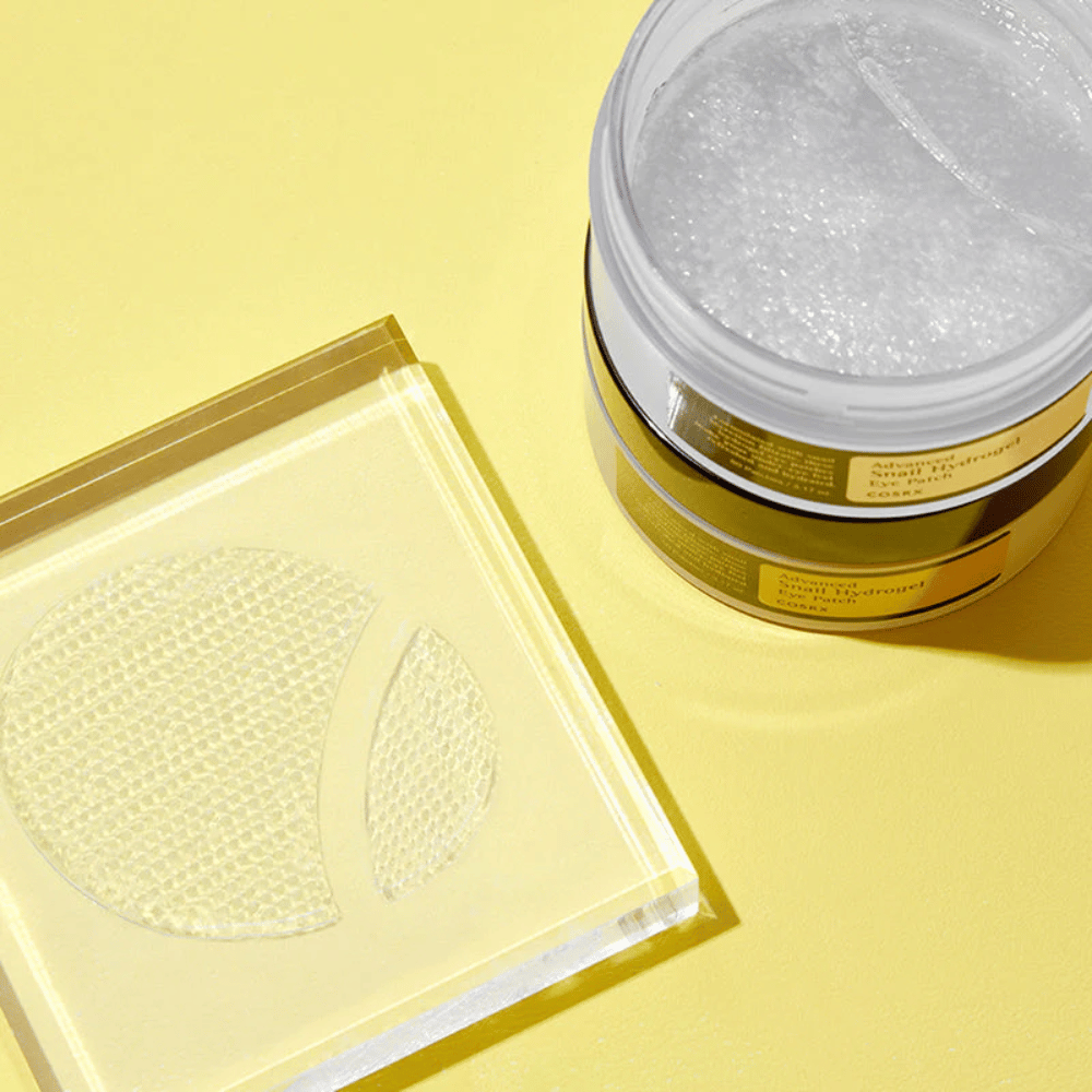 En åpen krukke COSRX Advanced Snail Hydrogel Eye Patch står på gul bakgrunn ved siden av en gjennomsiktig underlag hvor to øyenlapper ligger plassert.