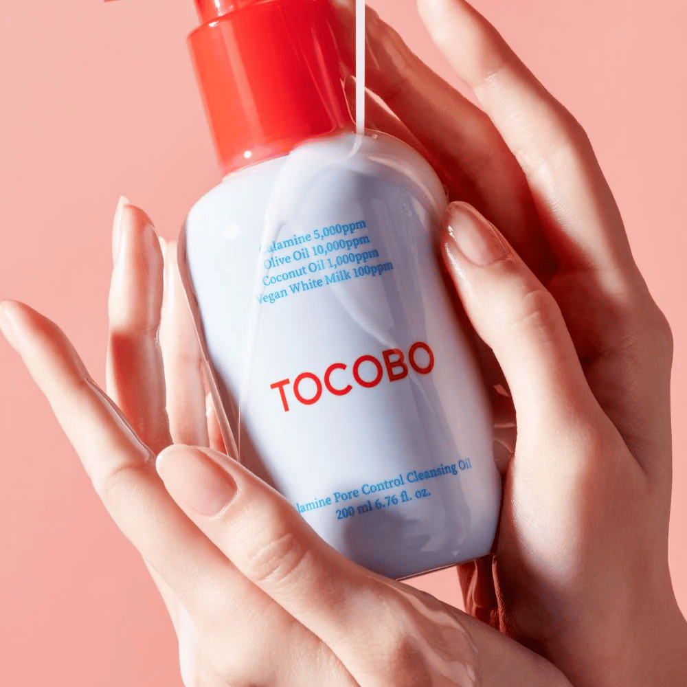TOCOBO Calamine Pore Control Cleansing Oil holdes i hender mot en rosa bakgrunn, flaskens røde pumpe og ingrediensliste er tydelig synlig.