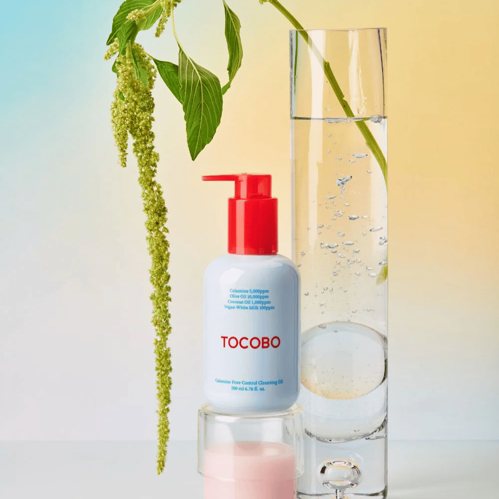 TOCOBO Calamine Pore Control Cleansing Oil med rød pumpe står ved siden av et høyt glassrør fylt med vann og en grønn plante, mot en lys pastellfarget bakgrunn.