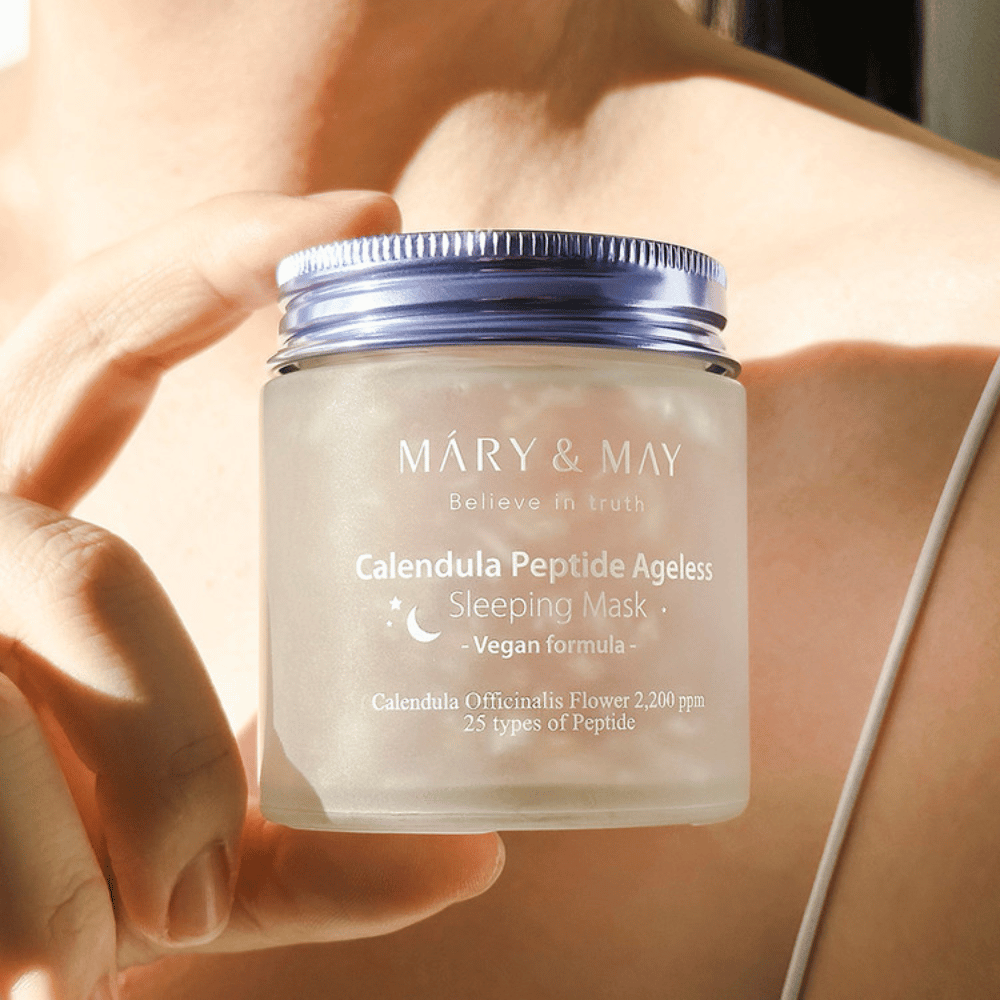 En kvinne holder en boks med MARY & MAY Calendula Peptide Ageless Sleeping Mask. Boksen er gjennomsiktig med et lilla lokk og teksten "Calendula Peptide Ageless Sleeping Mask" er tydelig synlig. Produktet er en vegansk sovemaske med calendula og peptider, beregnet på å gi huden en ungdommelig glød om natten.