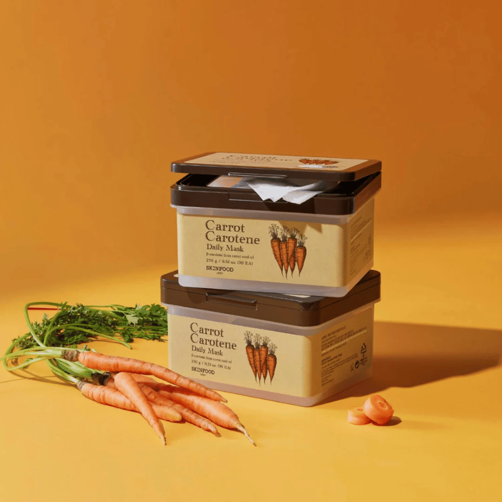 Bildet viser tre stablede pakker av "Carrot Carotene Daily Mask" fra SKINFOOD, plassert på en oransje bakgrunn sammen med ferske gulrøtter. Hver eske inneholder ansiktsmasker beriket med β-karoten fra gulrotfrøolje, designet for daglig bruk for å nære og pleie huden.