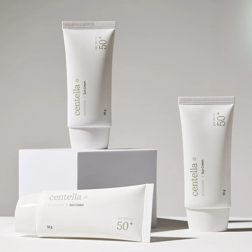 Tre pakker med Centella Sun Cream SPF 50+ PA++++ arrangert estetisk på hvite podier. To tuber står opp og en ligger ned, alle med hvit, minimalistisk design og grønne tekstdetaljer.